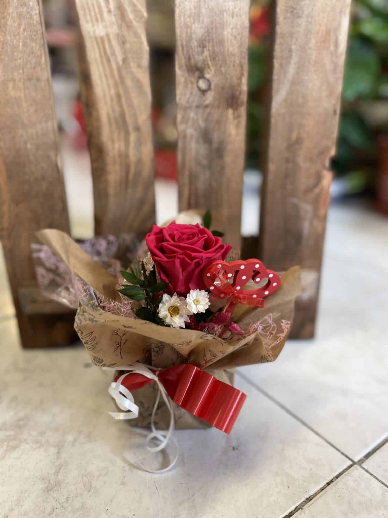 Rosa preservada regalo de Floristería Sayde, con una rosa roja premium, flores complementarias blancas, follaje verde, y decoración artesanal en papel kraft, adornada con un lazo rojo y blanco, ideal para sorprender en ocasiones especiales como San Valentín