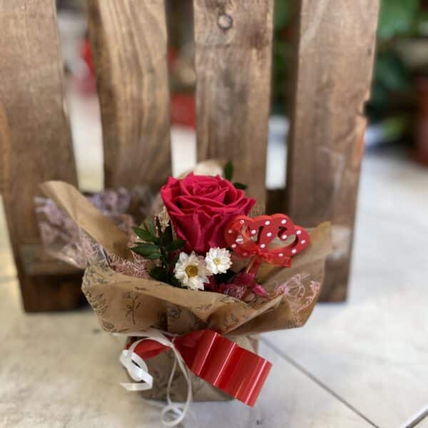 Rosa preservada regalo de Floristería Sayde, con una rosa roja premium, flores complementarias blancas, follaje verde, y decoración artesanal en papel kraft, adornada con un lazo rojo y blanco, ideal para sorprender en ocasiones especiales como San Valentín